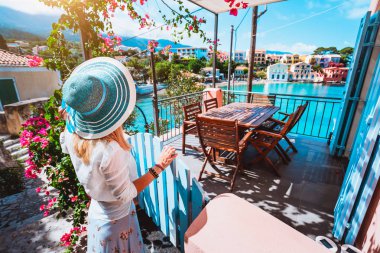 Assos köyün önünde rahat veranda kalıyor ve turkuaz renkli bay Akdeniz deniz ve Kefalonia, Yunanistan için güzel renkli evlerin hayran mavi güneş şapkalı kadın turist