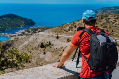 Male summer holiday vacation on Kefalonia Greece. Photographer with backpack enjoying capture of Mediterranean village Assos from top view point. Camera on tripod clipart