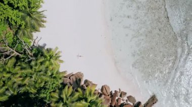 4k havadan görünümü dikey hareket aşağı kız beyaz bir kumlu sahilde döşeme kristal berraklığında turkuaz sığ okyanus Lagoon su ve muhteşem granit kayalar tropikal La Digue adada çevrili