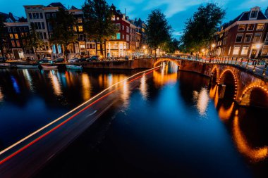 Amsterdam akşamı şehir merkezinde aydınlandı. Leidsegracht ve Keizersgracht kanallarındaki turistik tekne ışık yolları ve köprü yansımaları. Uzun pozlu çekim. Şehir gezisi konsepti