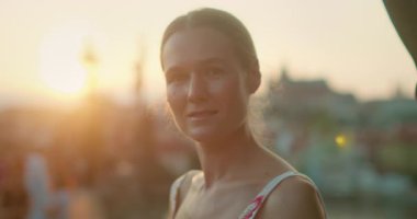 Prag 'da yaz aylarında bir köprüde gün batımında yumuşak bir gülümsemeyle kameraya bakan bir kadın..