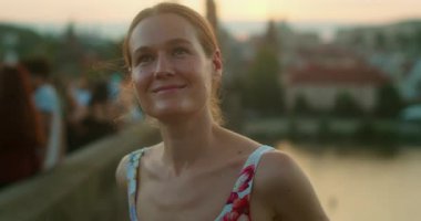 Çiçekli elbiseli genç bir kadın günbatımında tarihi bir Avrupa köprüsünde dikilirken omzunun üzerinden bakar ve gülümser..