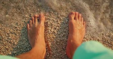 Çakıl taşlı bir sahilde duran çıplak ayaklı bir adamın altın saatinde hafif deniz dalgalarıyla yıkanan.