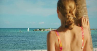 Güneşli sahilde palmiye ağacında dinlenirken okyanusa bakan bir kadın..