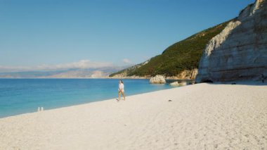 Kefalonya 'daki kayalıkların yanında, uzak beyaz çakıl kumsalı boyunca yürüyen kişi.