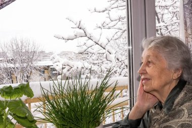 Yaşlı yalnız kadın evinin penceresinin yanında oturuyor ve kışın bakıp hayal kuruyor.