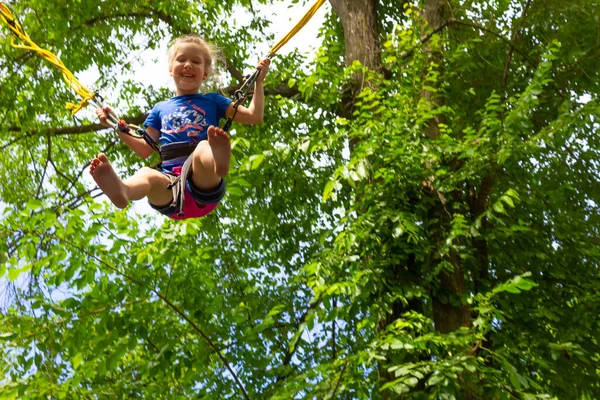 緑の木に対して公園でトランポリンでバンジーと一緒にジャンプ笑顔幸せな女の子 — ストック写真