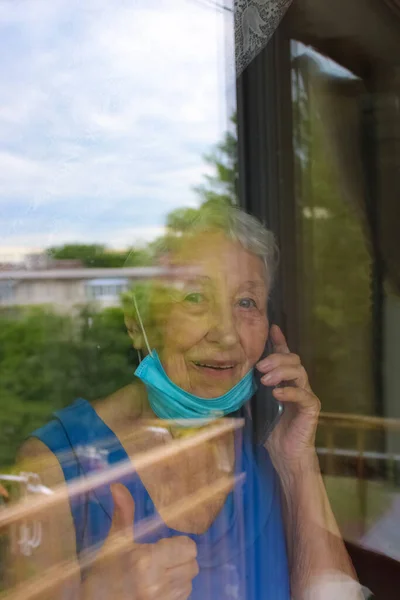 The Covid-19, health, safety and pandemic concept - senior old lonely woman wearing protective medical mask sitting near the window at home for protection from virus with mobile phone