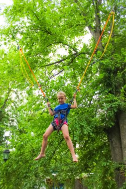 Gülümseyen mutlu kız yeşil ağaçlara karşı trambolinde bungee ile atlıyor.