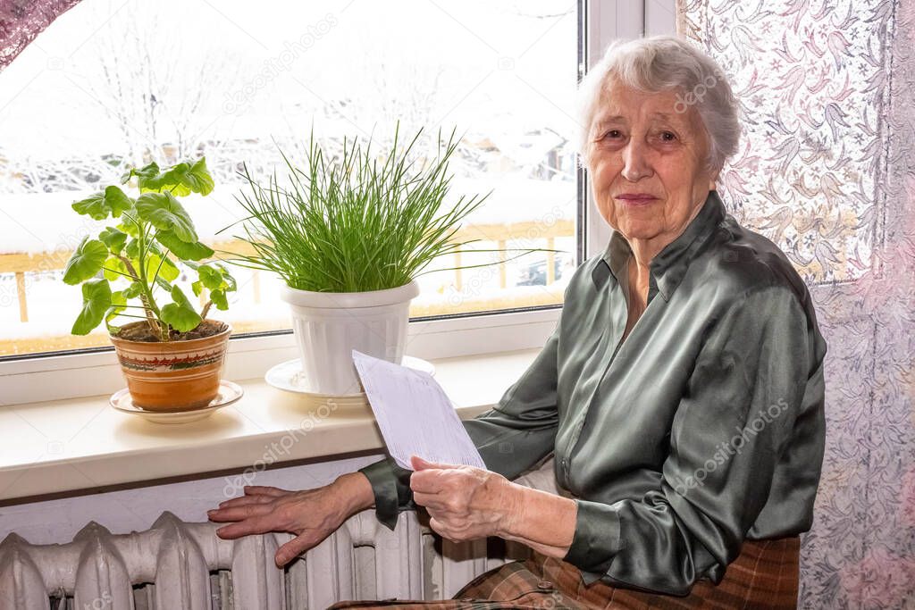 The sad senior woman holding gas bill in front of heating radiator. Payment for heating in winter.