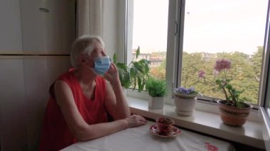 Covid-19, sağlık, güvenlik ve salgın konsepti - koruyucu maske takan yaşlı, yalnız bir kadın virüsten korunmak için evdeki pencerenin yanında oturuyor.