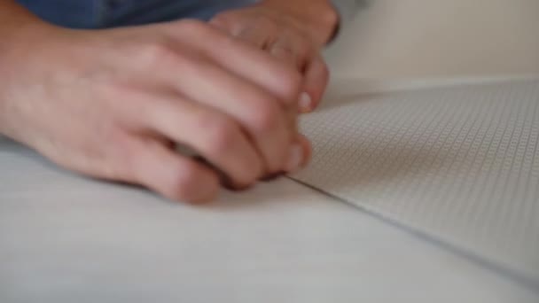 Primer plano manos masculinas preparando nuevo rollo de papel pintado doblado con cuchillo de papelería en la mesa de interior — Vídeos de Stock