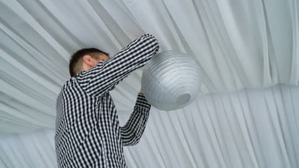 Man hanging colorful white pink gray paper lanterns to ceiling. — Stock Video