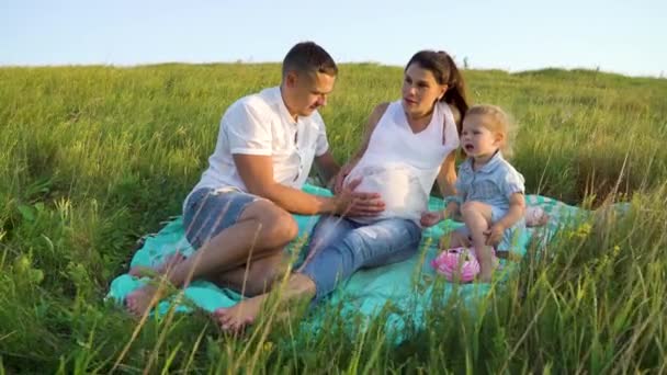 Pregnant couple with toddler daughter have leisure time outdoorsin grass field on blanket — Stock Video