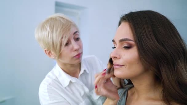 Zbliżenie wizażystka blondynka robi profesjonalny makijaż dla Brunetki słodkie woman w salonie kosmetycznym — Wideo stockowe