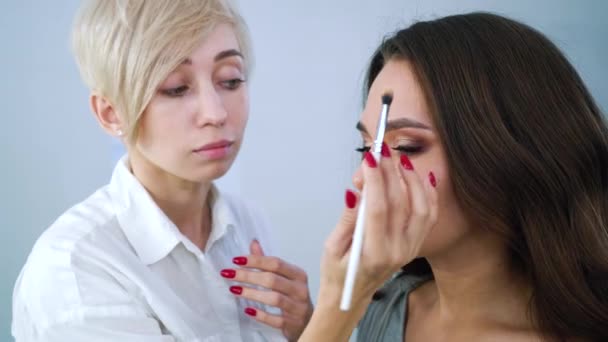 Primer plano artista de maquillaje femenino aplicando cosméticos en la cara hermosa mujer con cepillo en el salón de belleza — Vídeos de Stock