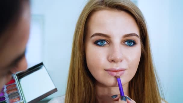 Close up artista de maquiagem feminina fazendo maquiagem profissional para jovem mulher de cabelo vermelho no salão de beleza — Vídeo de Stock