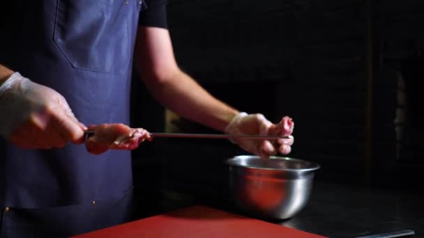 Молодой шеф-повар в фартуке и перчатках кладет куски маринованного мяса на шампур — стоковое видео
