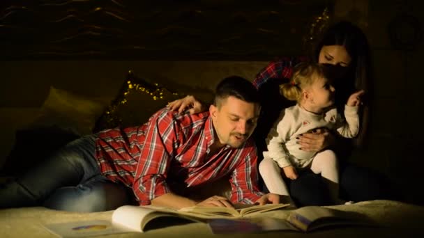 Счастливая семья с маленькой дочкой, читающей детские книги в постели по ночам — стоковое видео