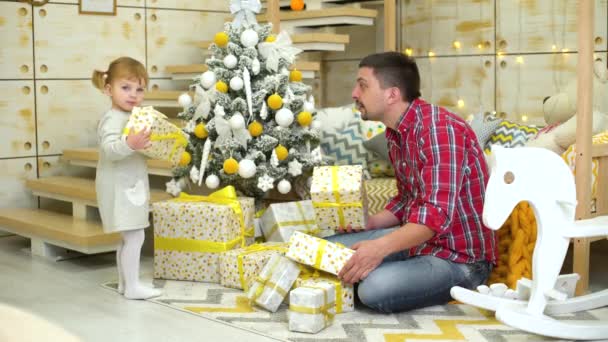 Kleines Mädchen mit Vater schüttelt Geschenkboxen in der Nähe des Weihnachtsbaums zu Hause — Stockvideo