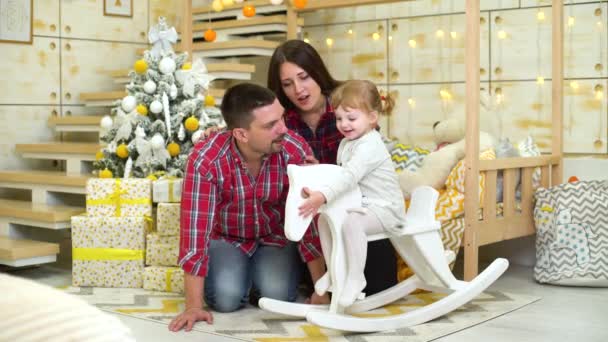 Niña columpios en balanceo caballo al lado de sus padres cerca de árbol de Navidad — Vídeos de Stock