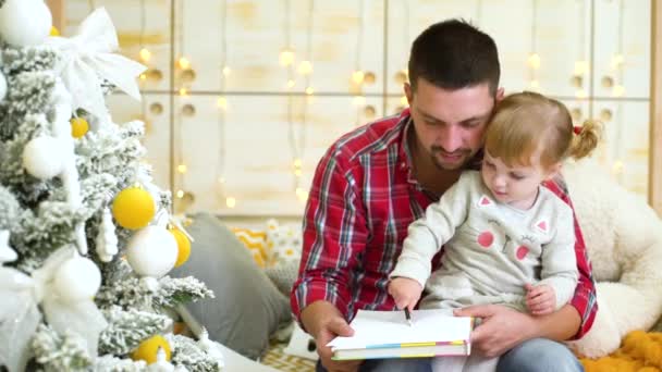 Niña pequeña sentada en su regazo padre y dibuja con lápiz cerca del árbol de Navidad — Vídeos de Stock