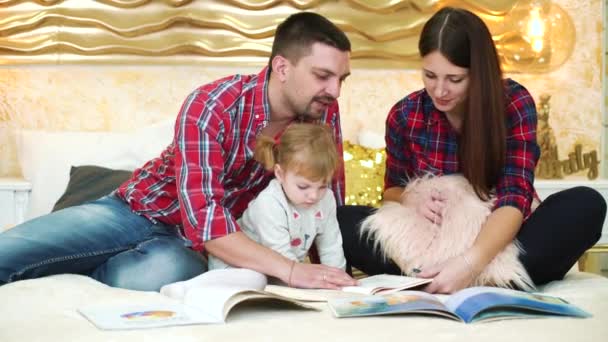 Padres jóvenes con una hija pequeña leyendo libros para niños en la cama en casa — Vídeo de stock