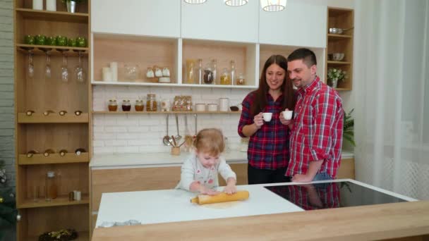 Schattig klein meisje rollen deeg, terwijl haar gelukkige ouders koffie in de keuken drinken — Stockvideo