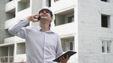 İnşaat sitesi mühendis cep telefonu kontrol üzerinde konuşurken tablet ile inşa teknoloji süreci. Kask içinde erkek mimar iş akışı açık havada denetler. Bina, sanayi, emniyet konsepti