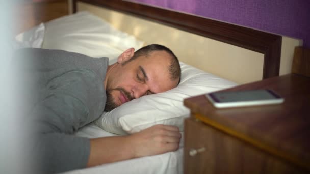 Молодой бородатый мужчина, спящий в постели, пробуждается сигналом тревоги на телефоне — стоковое видео