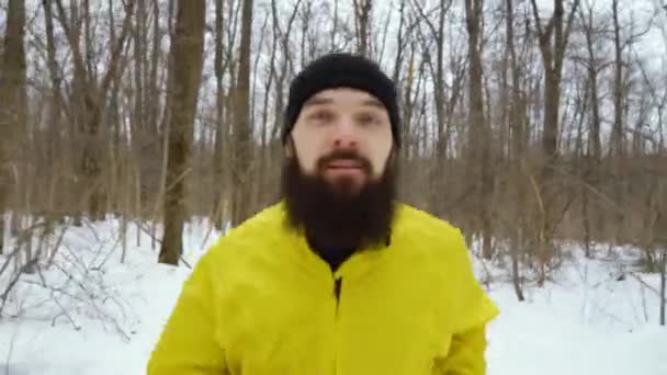 Крупный план бородатого спортсмена в желтом пальто, бегущего в снежном зимнем лесу — стоковое видео