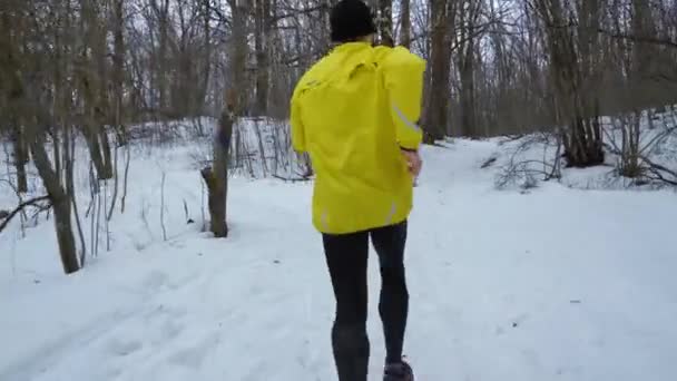 Tracking shot van sportman in gele jas de heuvel in winter bos aangelopen — Stockvideo