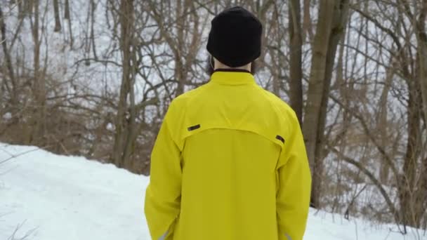 Vista trasera del hombre en abrigo deportivo amarillo de pie en la colina en el bosque de invierno — Vídeo de stock