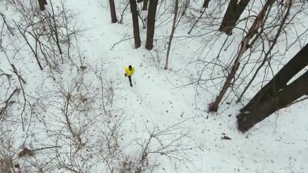 Plano de la cacerola aérea del deportista masculino en la capa amarilla corriendo en el bosque de invierno — Vídeo de stock