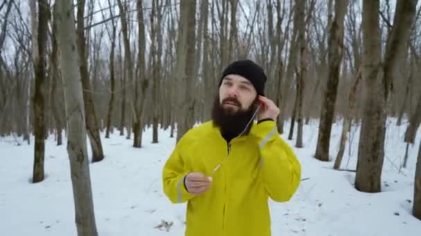 Бородатый спортивный человек надевает наушники и бегает в снежном зимнем лесу — стоковое видео