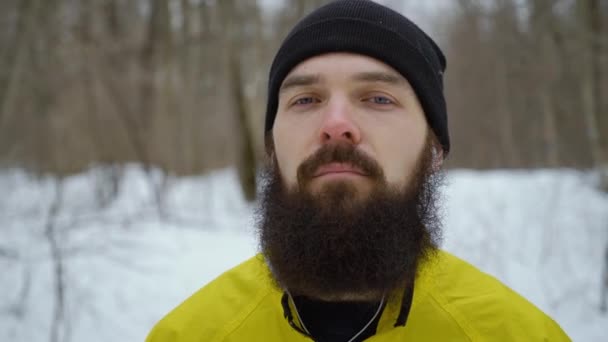 Крупный план хипстера с голубыми глазами и бородой, стоящего в зимнем лесу — стоковое видео