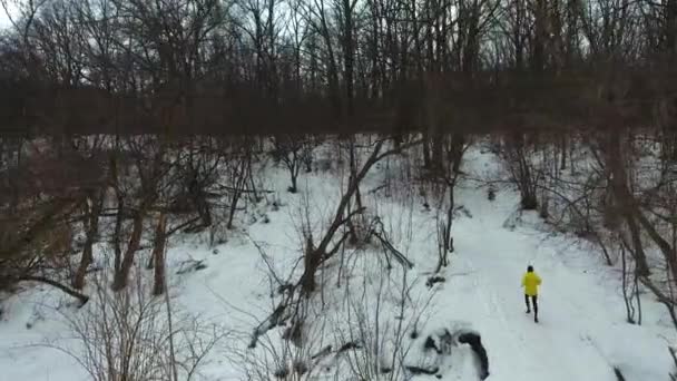 Tiro aéreo de deportista en abrigo amarillo corriendo por la colina en el bosque de invierno — Vídeos de Stock