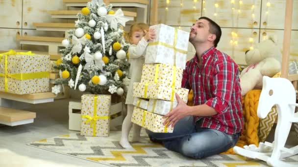 Маленька дівчинка з батьком розважається з подарунковими коробками біля ялинки вдома — стокове відео