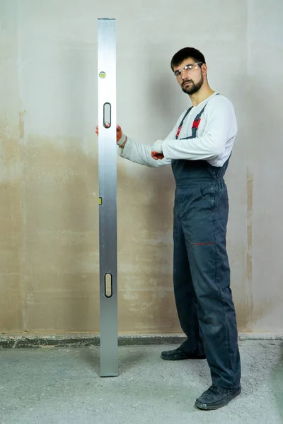 Young bearded builder stands near empty wall with construction ruler — 스톡 사진