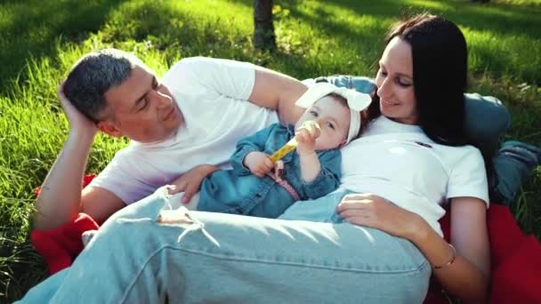 Любляча сім'я з дитиною на ковдрі на зеленій траві в літньому саду — стокове відео
