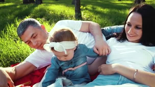 Lachende ouders met baby meisje ontspannen op deken op groen gras in Park — Stockvideo