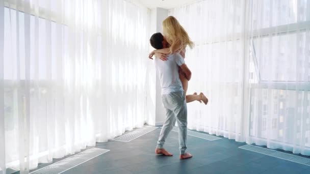 Jovem homem segurando mulher loira nos braços girando ao redor perto de grande janela francesa — Vídeo de Stock