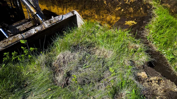 Vista recortada del cucharón excavador excavando en el campo herboso durante las obras de tierra —  Fotos de Stock
