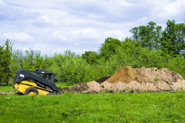 Grävmaskin grävande grop på gräsand fält med trädgård på bakgrunden — Stockfoto