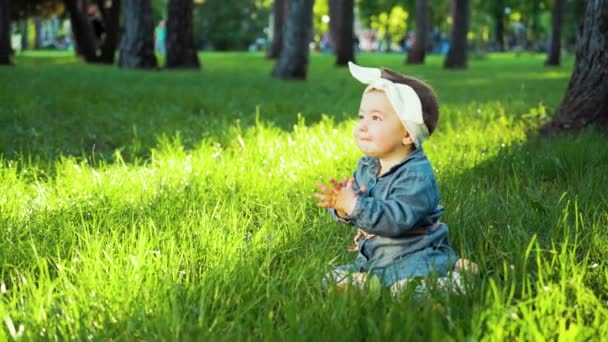 Gadis manis dengan celana jeans dan pita rambut duduk di rumput hijau di taman yang cerah — Stok Video