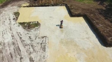 Titreşim plakalı bir adamın temel çukuruna kum fırlatırken havadan çekilmiş bir görüntüsü.
