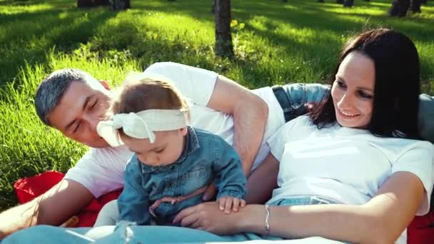 Весёлая девочка с родителями наслаждается солнечной погодой, лежащей на одеяле в парке — стоковое видео