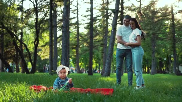 Мила дівчинка сидить на зеленій траві і люблячі батьки дивляться на неї ззаду — стокове відео