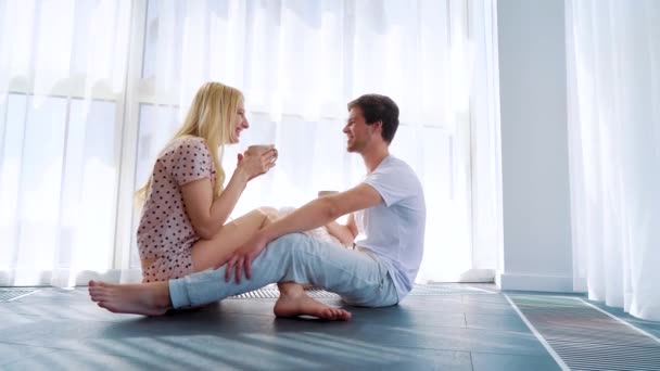 Boldog mosolygó pár szerelmes ülve a padlón, és iszik reggeli kávét — Stock videók