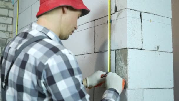 Mann in Arbeitskleidung und roter Mütze befestigt Metallschiene mit Klemmen an Blockwand — Stockvideo